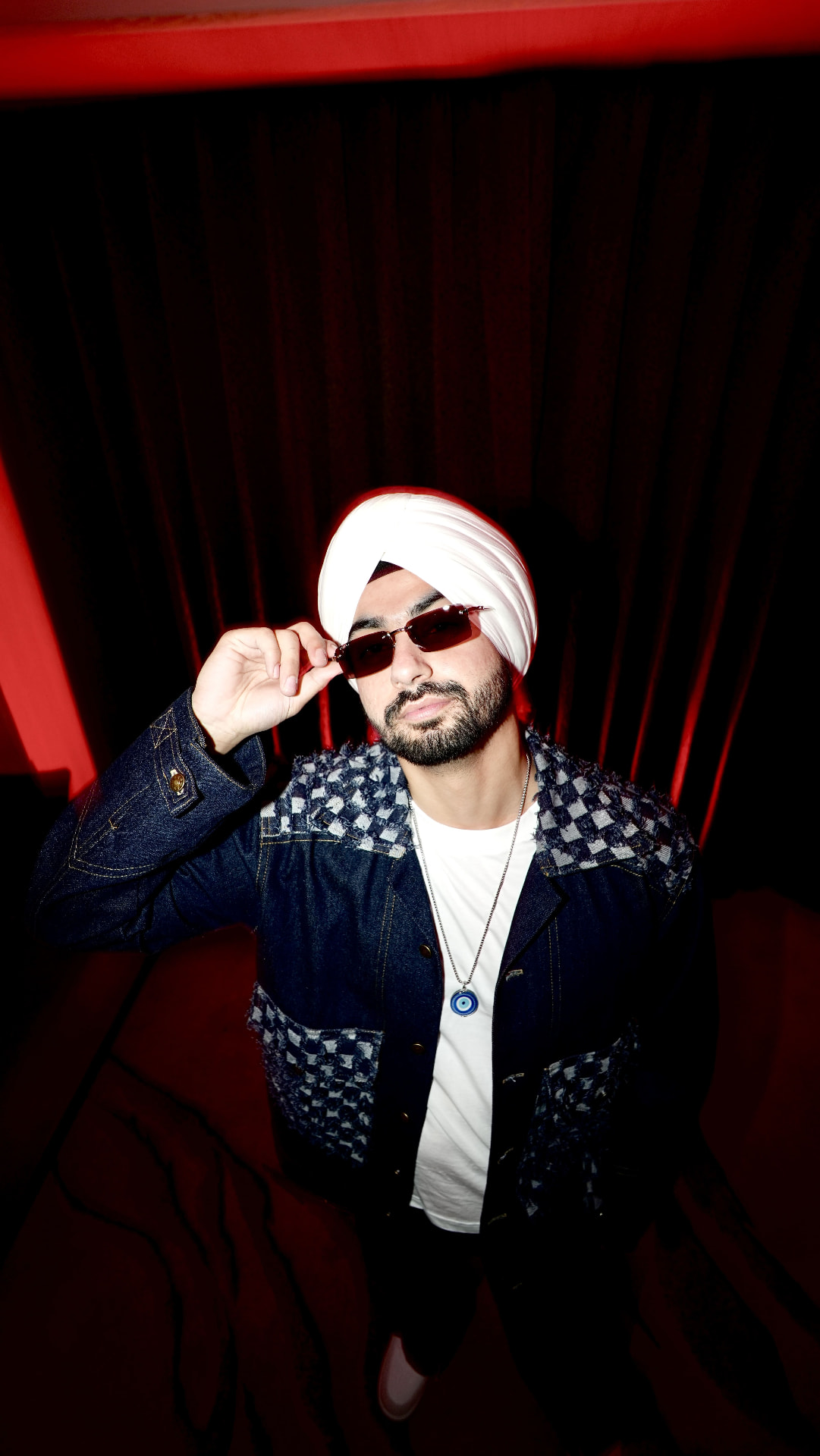 Sahib in a white turban and denim jacket lowering his sunglasses against a red curtain backdrop.