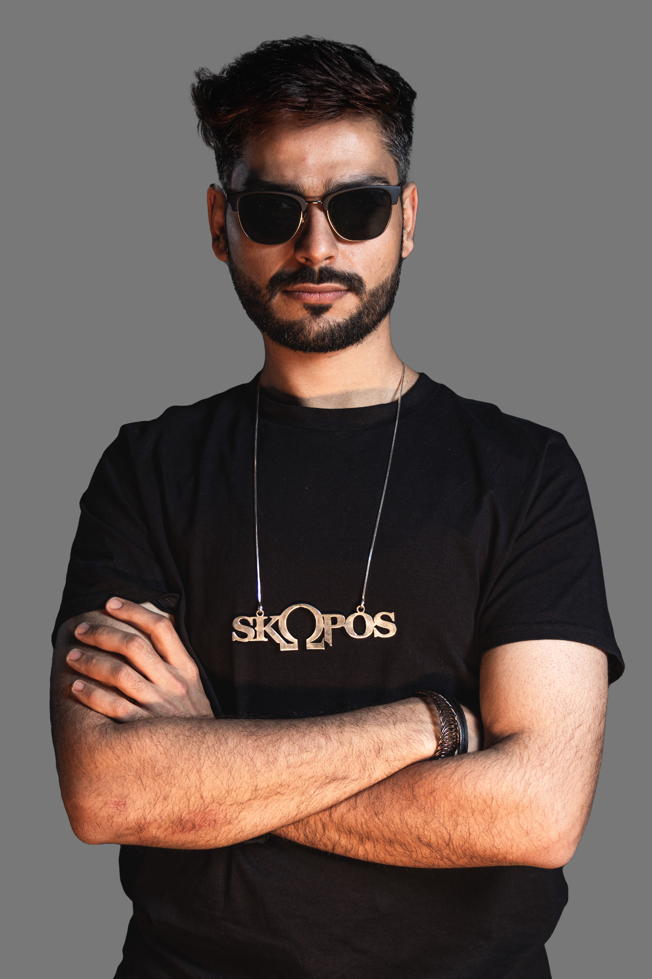 Skopòs in a black tee with the signature SKΩPOS chain, arms crossed.