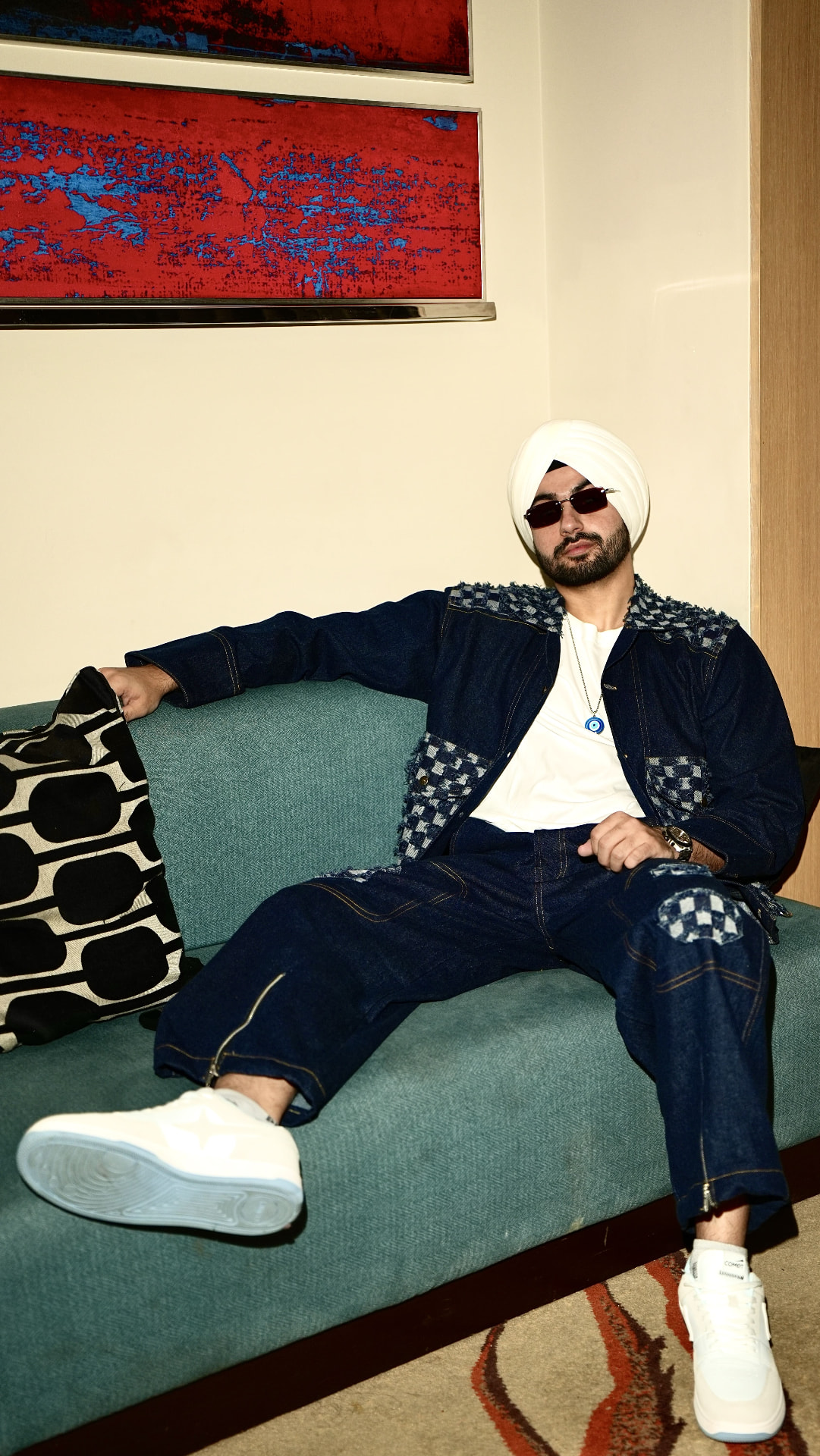 Sahib reclining on a teal sofa in a denim two-piece with red-and-blue artwork behind him.
