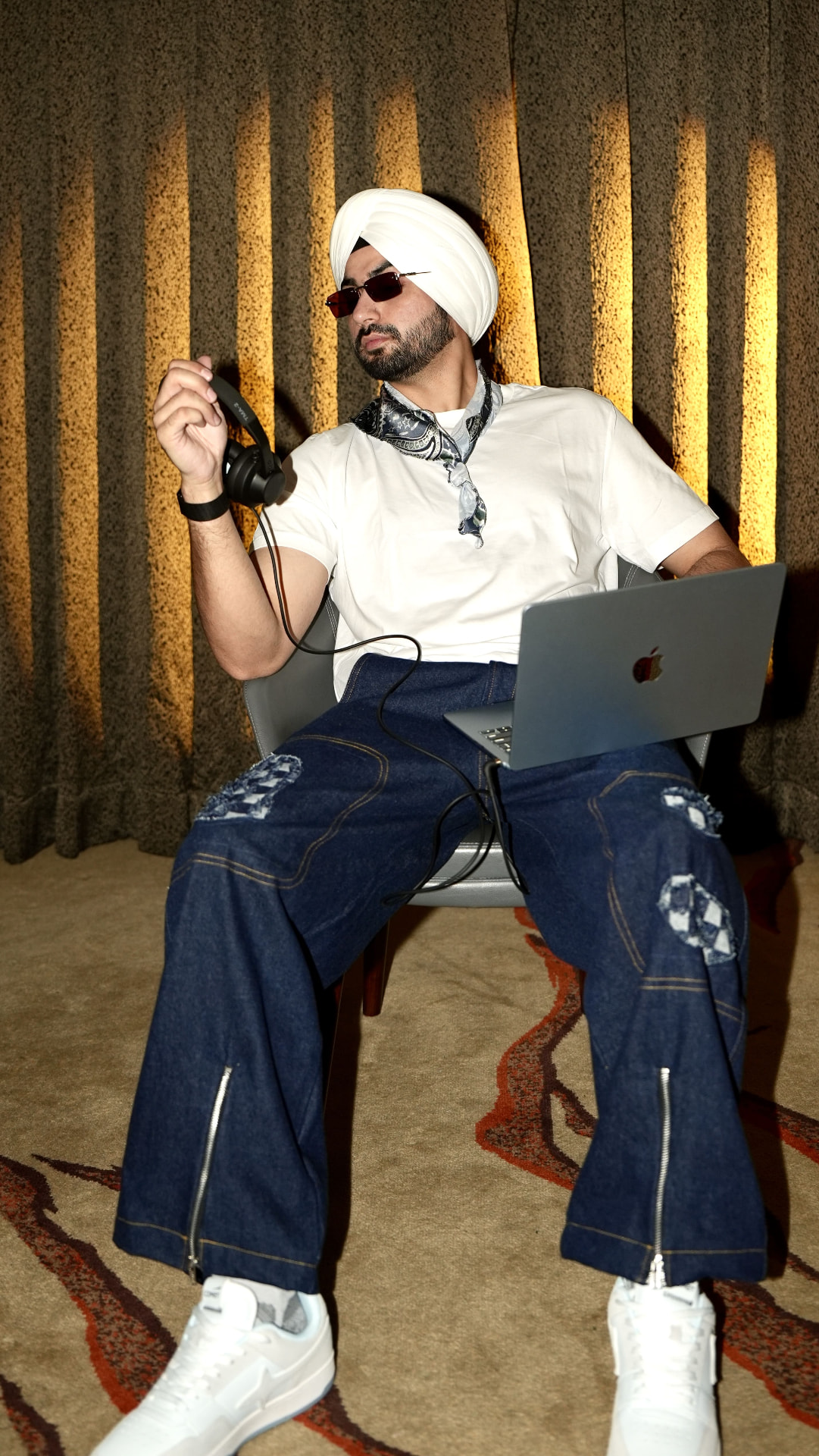 Sahib holding headphones with a laptop on his lap in a warmly lit lounge setting.
