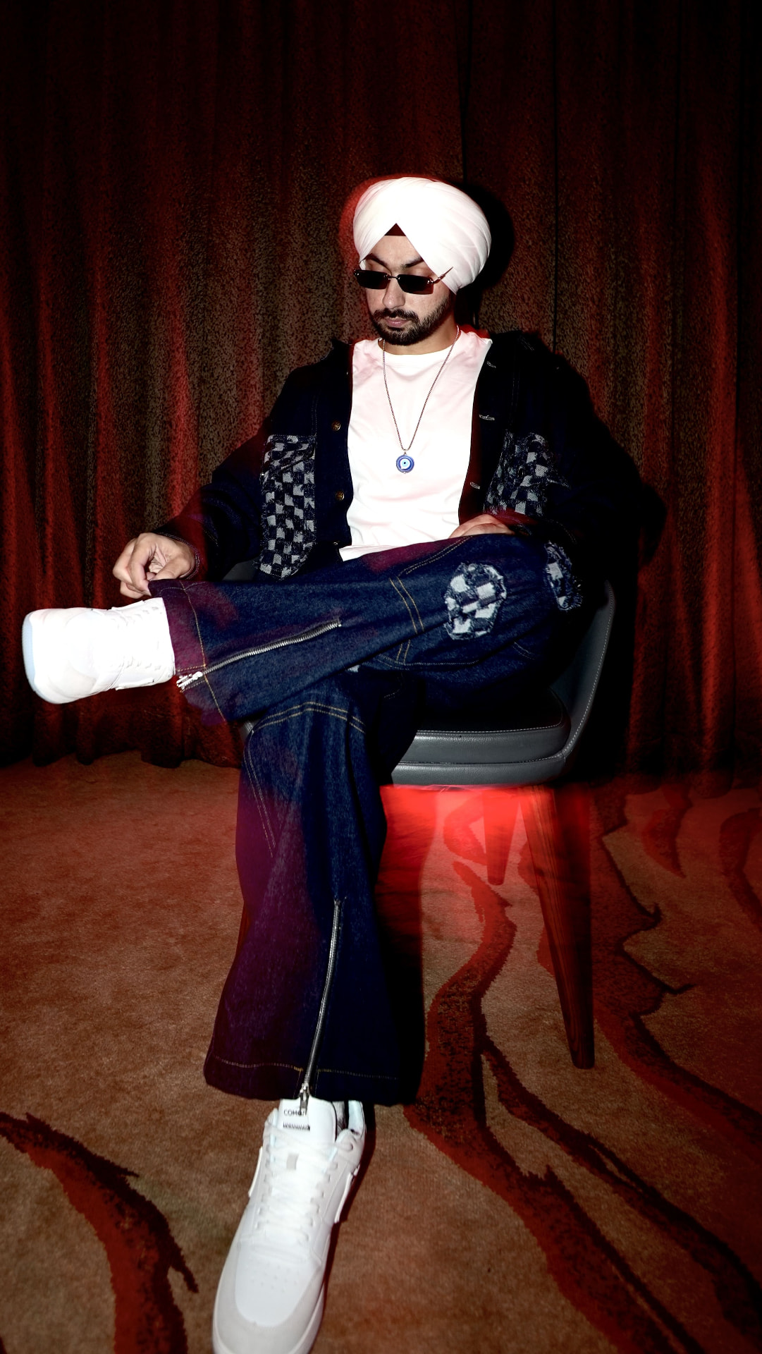 Sahib seated on a chair in a denim outfit and white sneakers under moody red lighting.