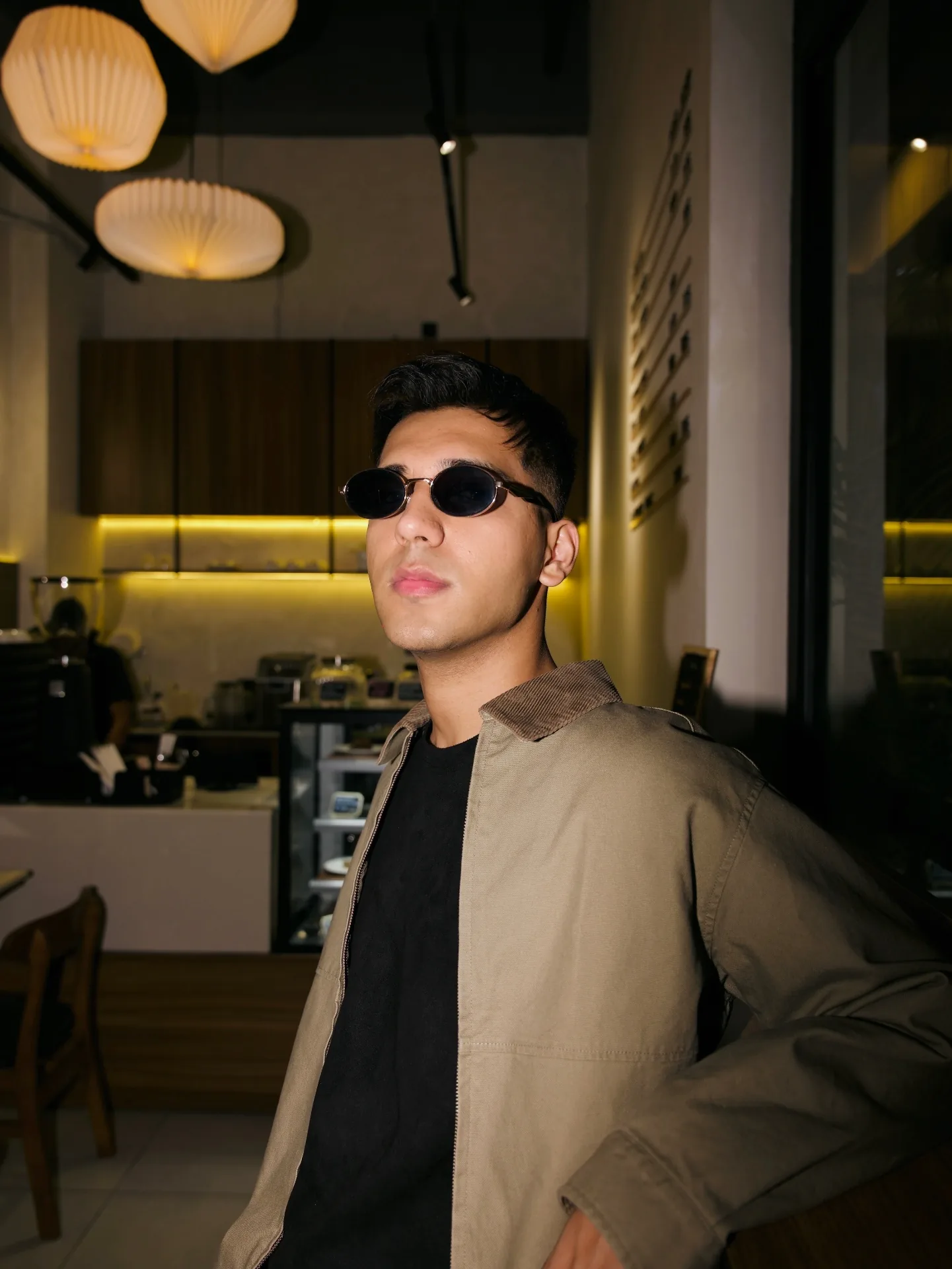 Exbow in shades and a khaki jacket inside a low-lit café.